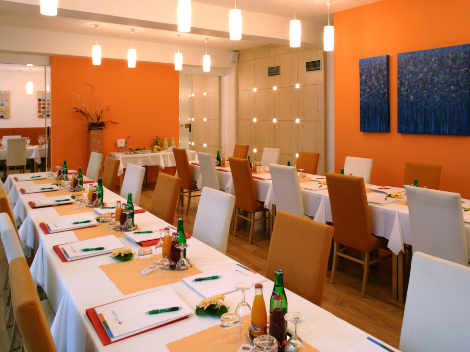 Restaurant dining room with long white-table banquet setup, orange accent walls, and modern pendant lights above.