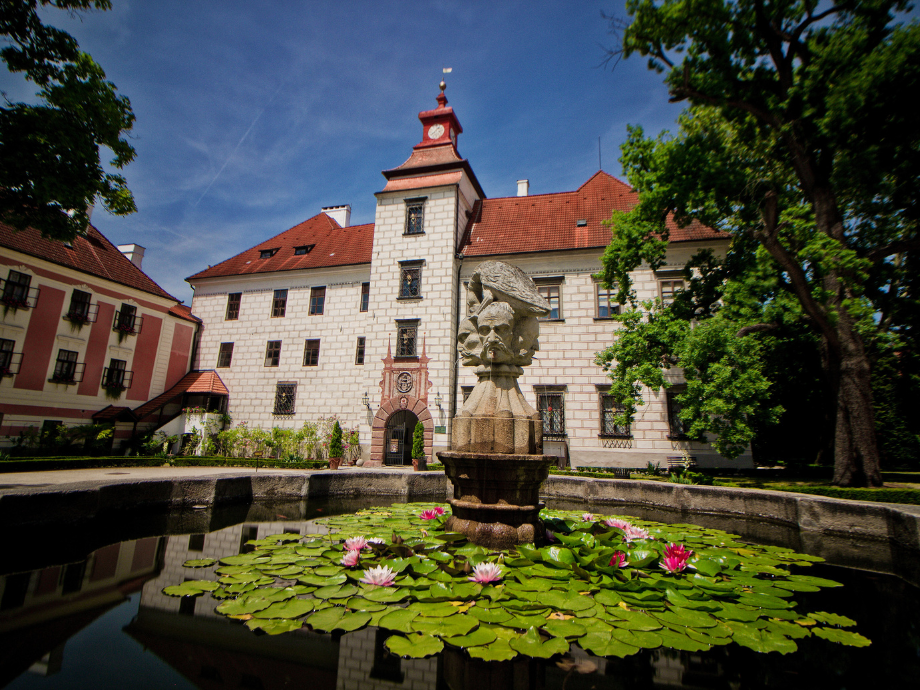 Stone courtyard with a fountain surrounded by lily pads and pink water lilies, a historic white stone building with a red-tiled roof in the background.