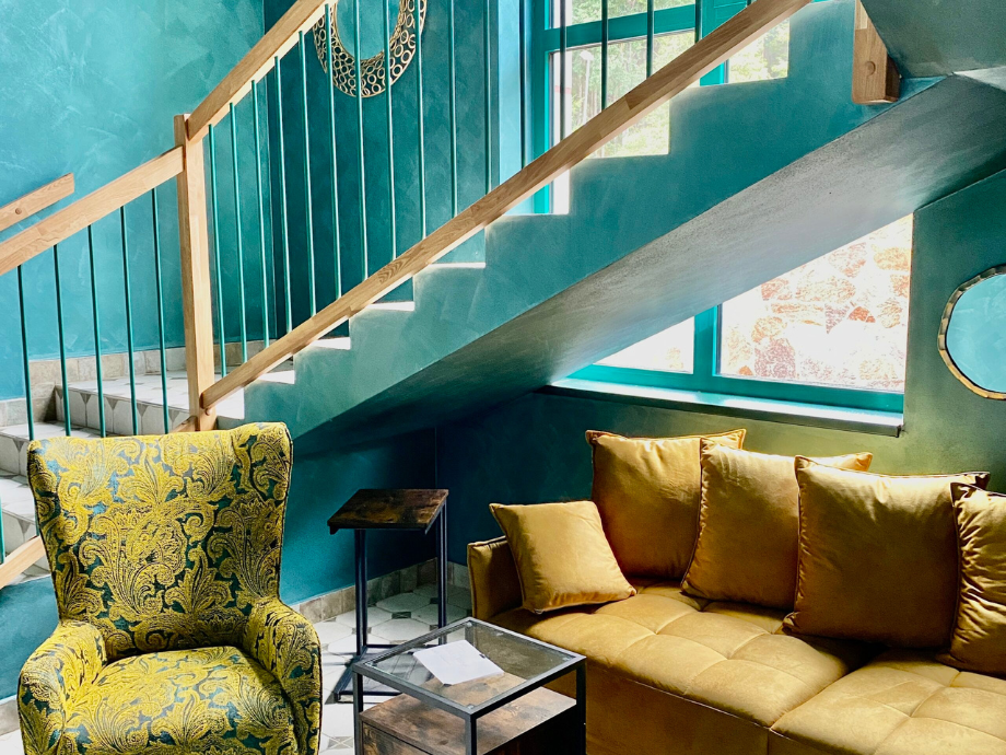 Teal living room under a staircase with a mustard-yellow sofa and a patterned green armchair, paired with a glass coffee table and a small side table by a large window letting in light.