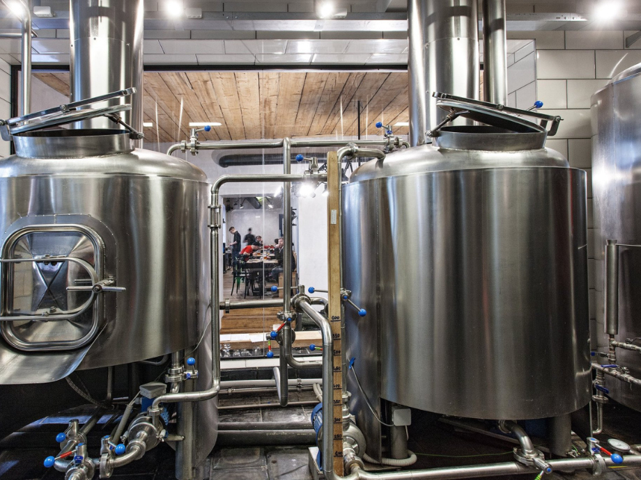 Two tall stainless steel fermentation tanks with a network of pipes in a brewery; a group of people sits at a table in the background.