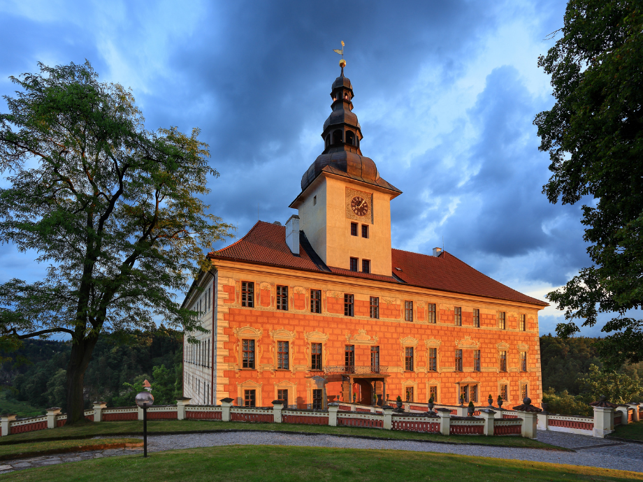 Historic brick castle with a tall clock tower bathed in warm sunset light, surrounded by trees and a fenced courtyard.