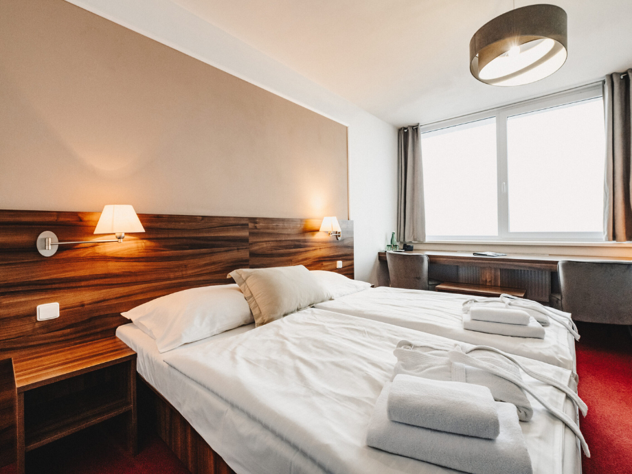 Hotel room with a large bed, wood headboard, wall lamps, and a windowed workspace by curtains.
