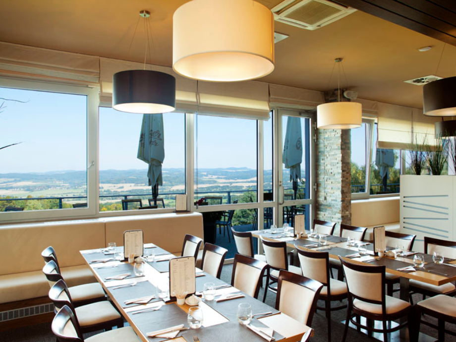 Bright dining area in a restaurant with large windows and a scenic landscape outside; tables are set with glasses, plates, and cutlery under large pendant lights.