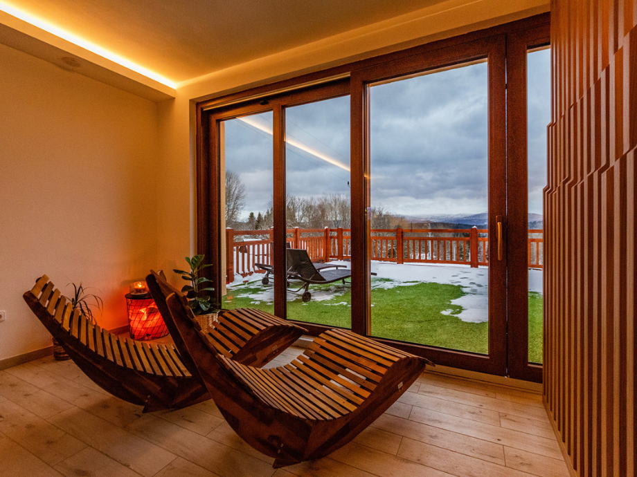 Indoor relaxation area with two curved wooden lounge chairs, a red lantern, and a potted plant, seen through large glass doors to a snowy balcony.