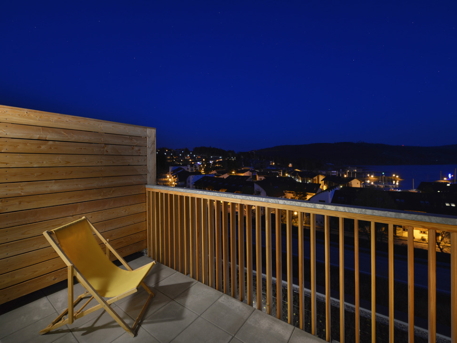 Nighttime balcony scene with a wooden wall, a yellow lounge chair, and a railing overlooking a lit coastal town and harbor below.