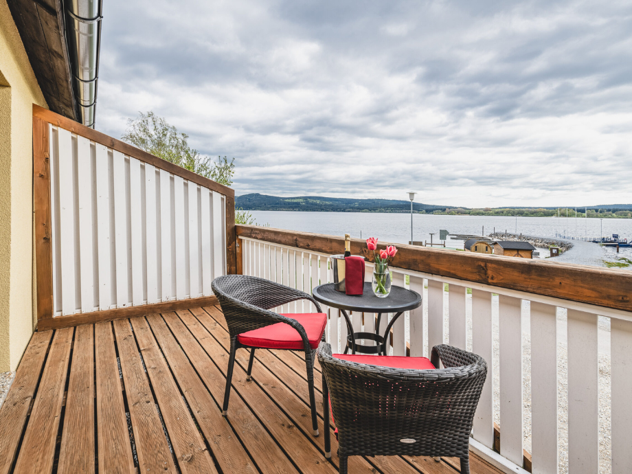 Balcony with wicker table and two chairs on wooden floor, overlooking a calm lake and boats.