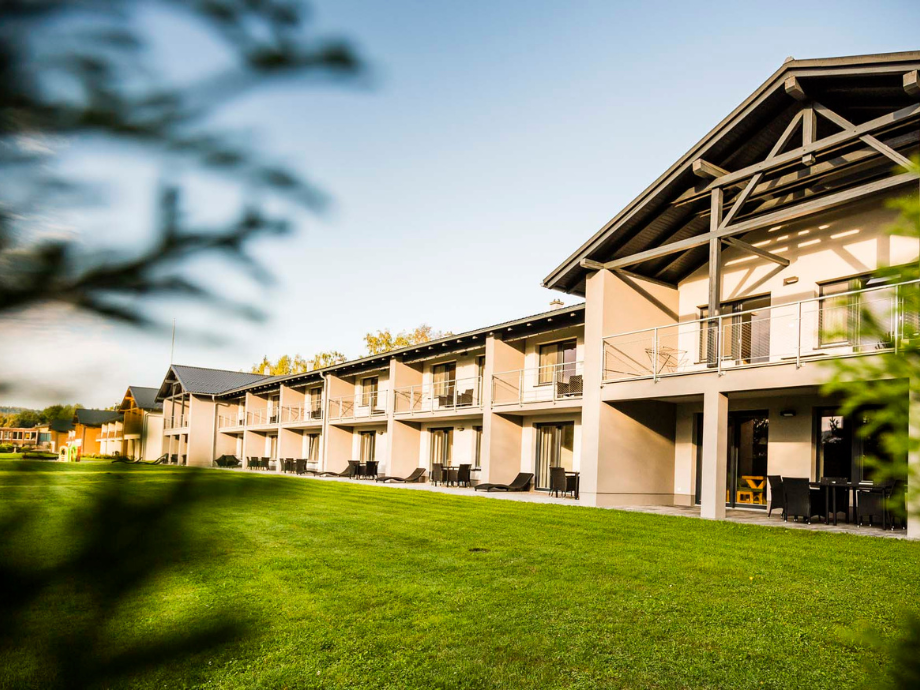 Row of modern townhouse-style hotel units with balconies along a green lawn under a clear blue sky, sunset light on the facade.