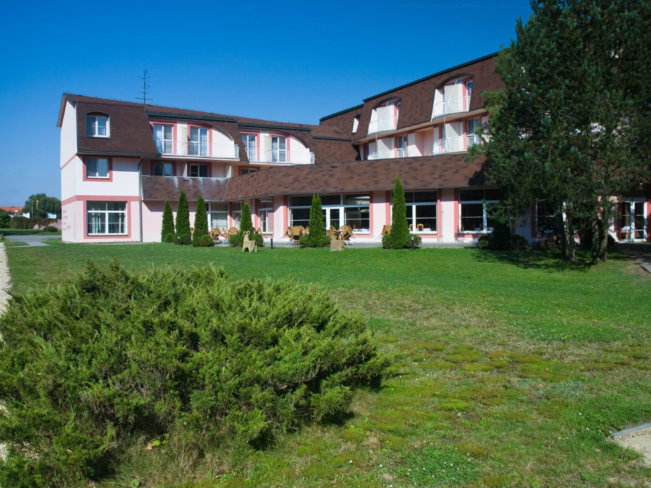 Two-story pink building with a brown mansard roof and balconies, set in a grassy courtyard under a blue sky.