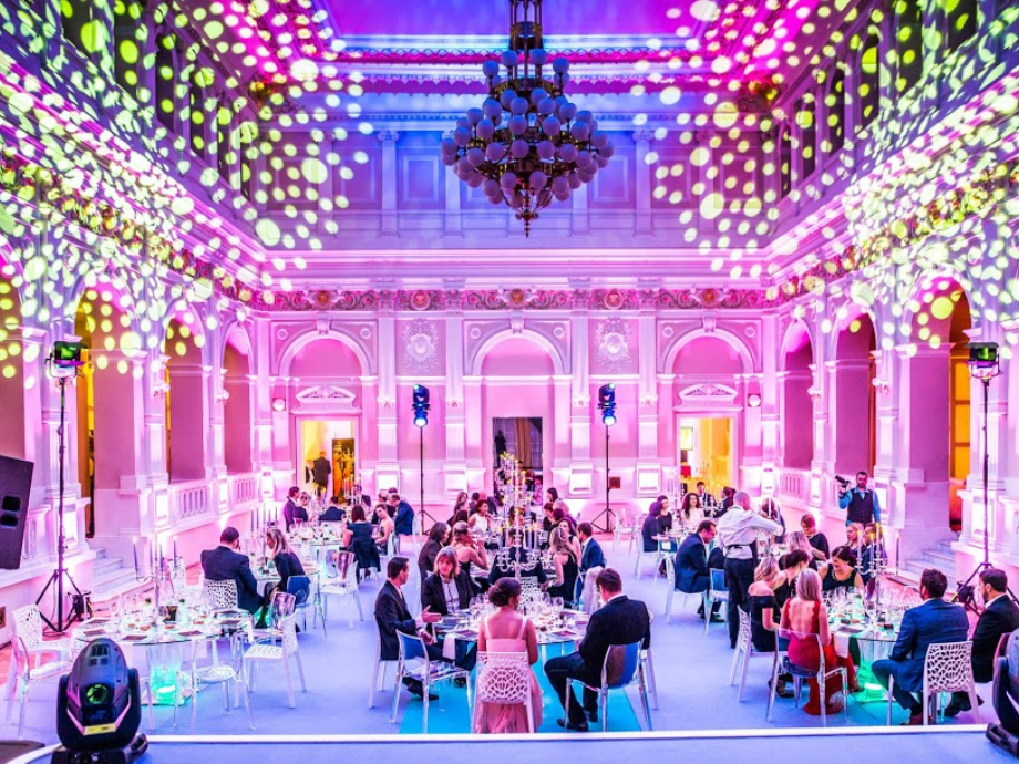 Grand ballroom with pink and purple lighting, a large chandelier, and guests seated at round tables for a formal banquet.