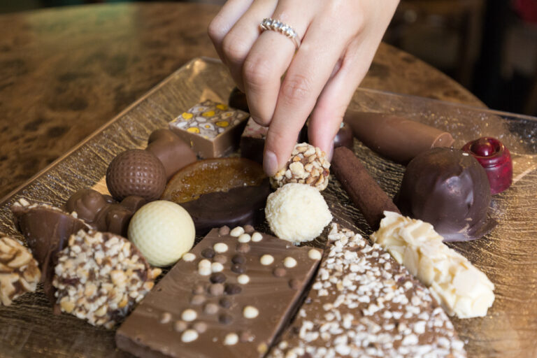 A hand with a ring picking a nut-coated chocolate truffle from a tray of assorted chocolates and confections.