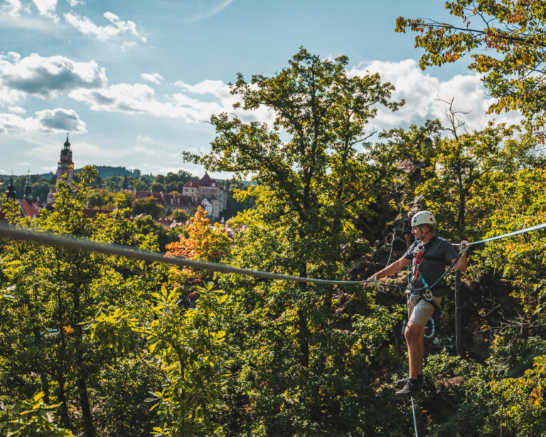 Man wearing a helmet and harness zip-lining across a forested hillside with a town and church spire in the distance.