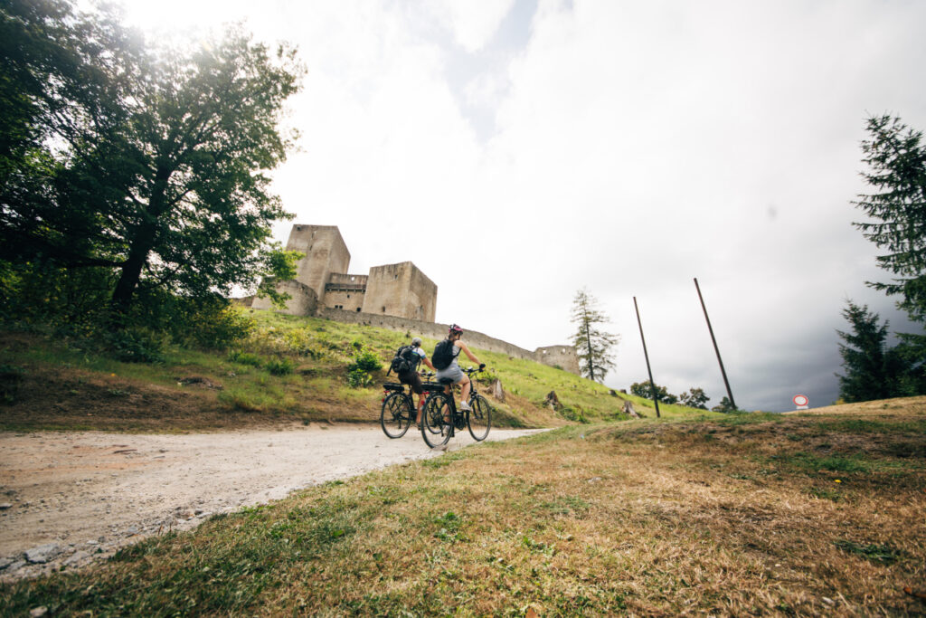 Two cyclists ride along a dirt path toward an ancient stone castle on a grassy hill.