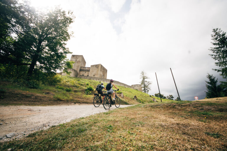 Two cyclists ride along a dirt path toward an ancient stone castle on a grassy hill.