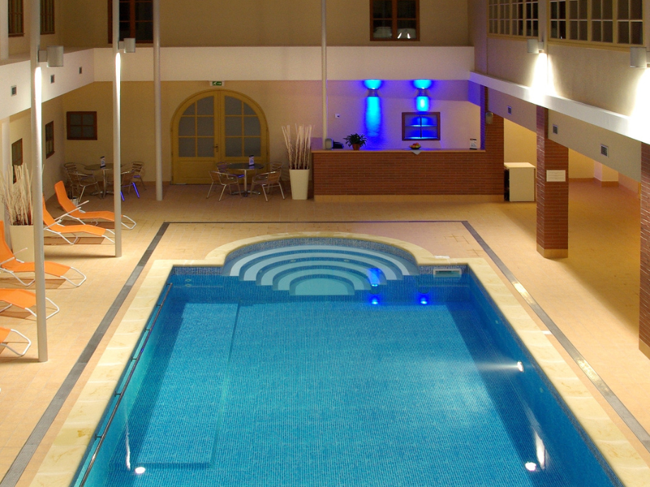 Indoor pool area with curved step入口 at one end, blue-tiled pool, beige surroundings, and orange lounge chairs along the left wall.
