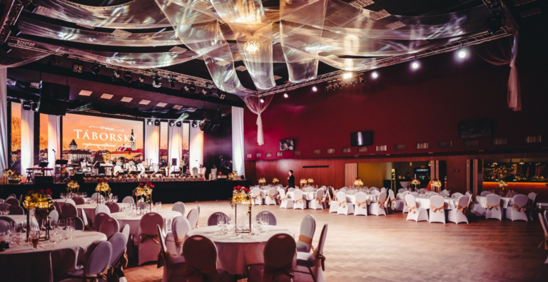 Elegant banquet hall with a live band on stage, velvet lighting, and draped ceiling decorations over round tables set with candles and gold bows.