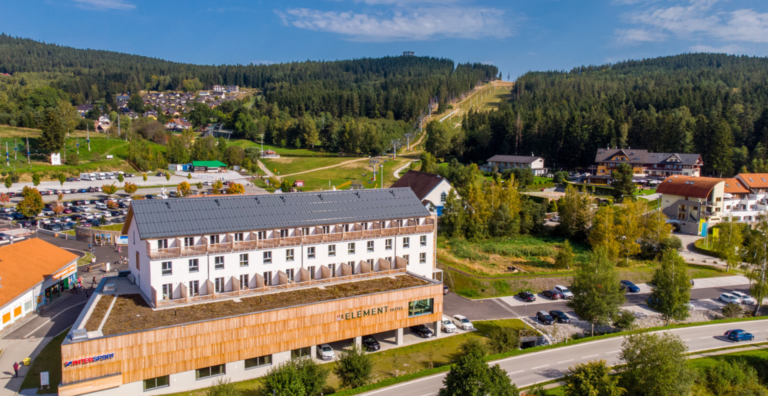 Aerial view of a mountain town with a large white hotel labeled 'Element' in the foreground and a hillside ski area in the background under a blue sky.