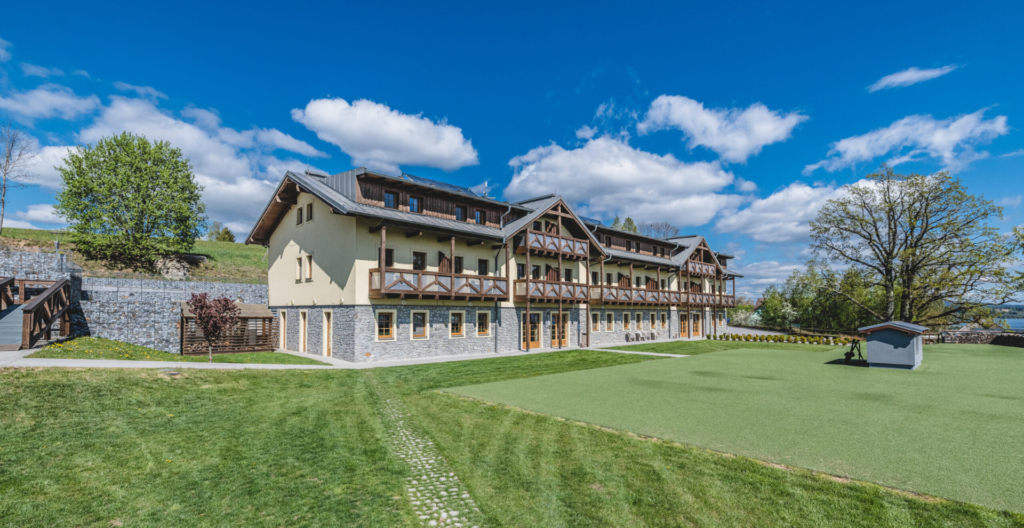 Large lodge-style building with stone foundation and wooden balconies set on a manicured lawn under a bright blue sky.