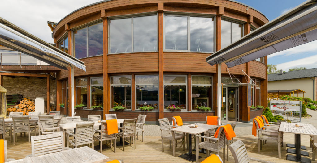 Outdoor café seating area with wooden tables and chairs, some orange cushions, in front of a curved wooden building with large glass windows and awnings.
