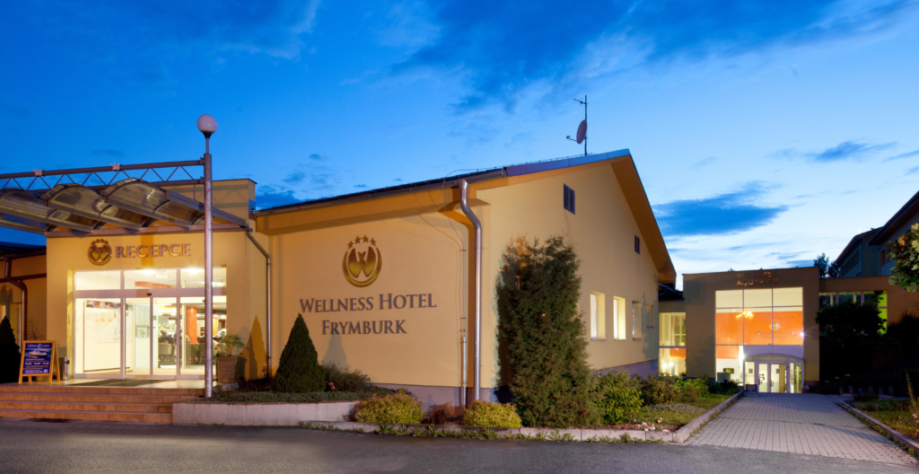 Exterior view of Wellness Hotel Frymburk at dusk, with illuminated reception entrance on the left and a large wall sign in the center.