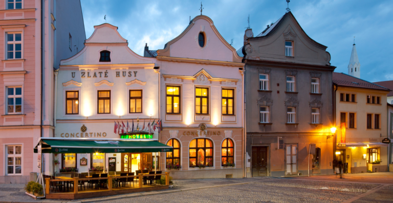 Evening street scene with historic buildings, outdoor cafe seating, and hotel signage reading 'Concertino' and 'U Zlaté husy'