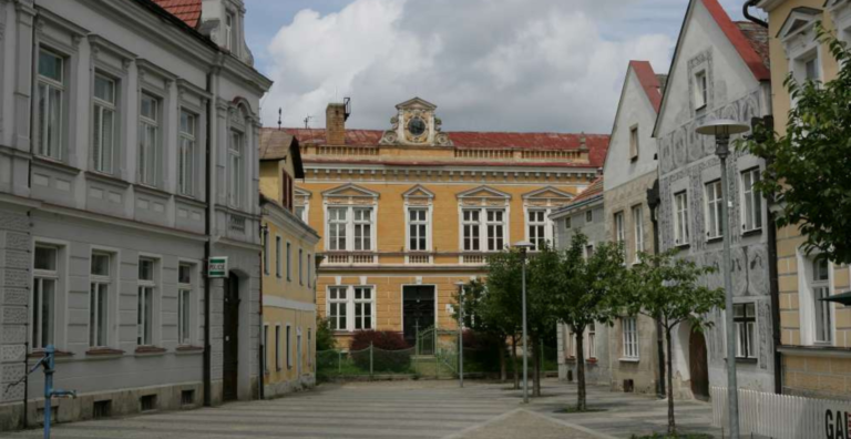 Historic yellow building with ornate facade at end of a tree-lined street, flanked by pale gray and patterned white buildings under a cloudy sky.