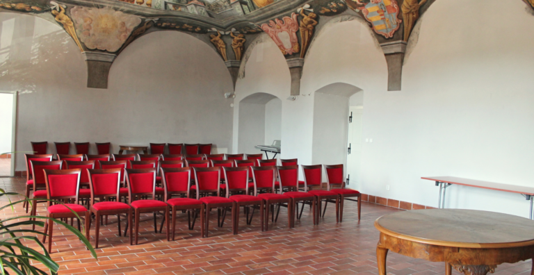 Historic hall set for an event: rows of red upholstered chairs facing a round stage, with decorative frescoed arches along the ceiling.
