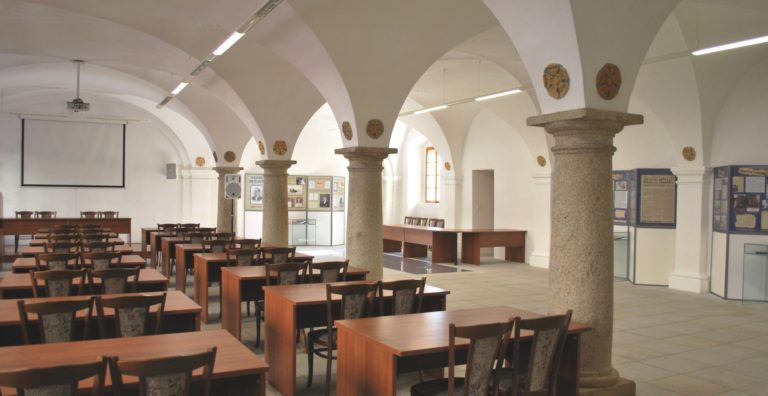 A large white lecture hall with tall arches and stone columns, rows of wooden desks and chairs, and a projector screen at the front.