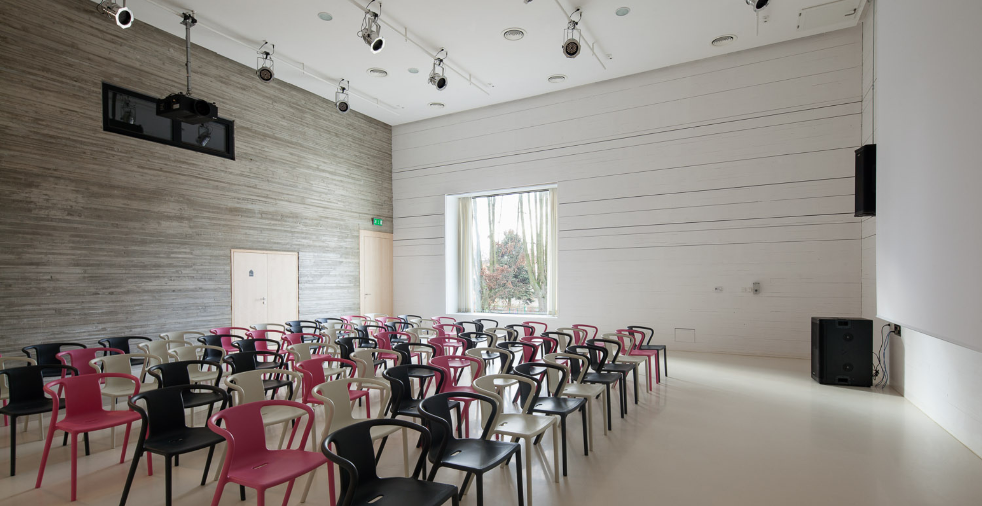 A bright, modern lecture room with rows of pink, black, and white chairs facing a blank wall; large window shows trees outside, ceiling lights and speakers above.