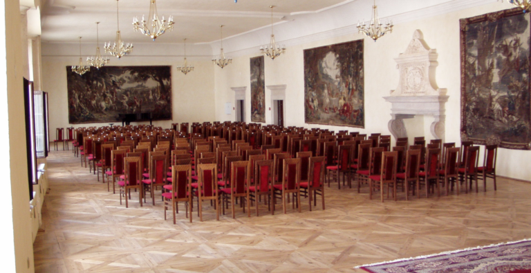 Lecture hall with rows of red-upholstered wooden chairs, chandeliers, and large wall tapestries on both sides.