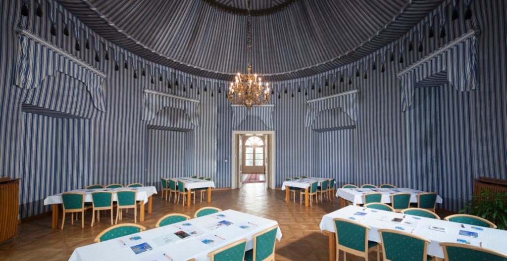 Formal dining hall with blue and white striped walls, a crystal chandelier, and round tables lined with green chairs.