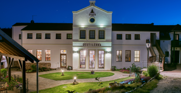 Front view of a white two-story distillery building at night, with a central gable that reads DESTILÉRKA and a circular window, illuminated entrance doors, and a landscaped courtyard.