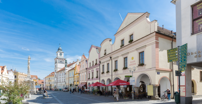 Sunny European street with pastel buildings, outdoor cafes under red umbrellas, and a clock tower in the background.