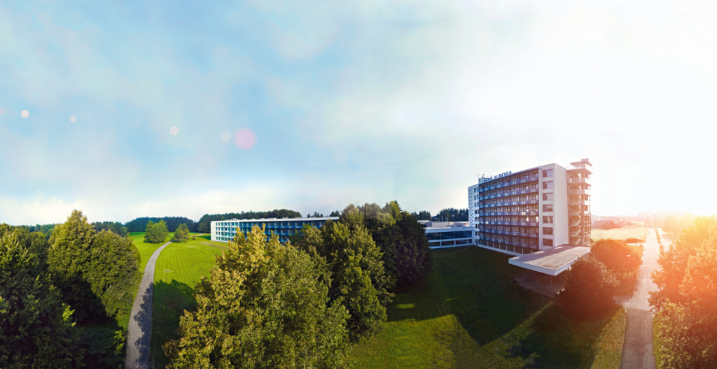 Aerial view of a modern hotel complex with white, multi-story buildings surrounded by green lawns and trees under a bright blue sky.