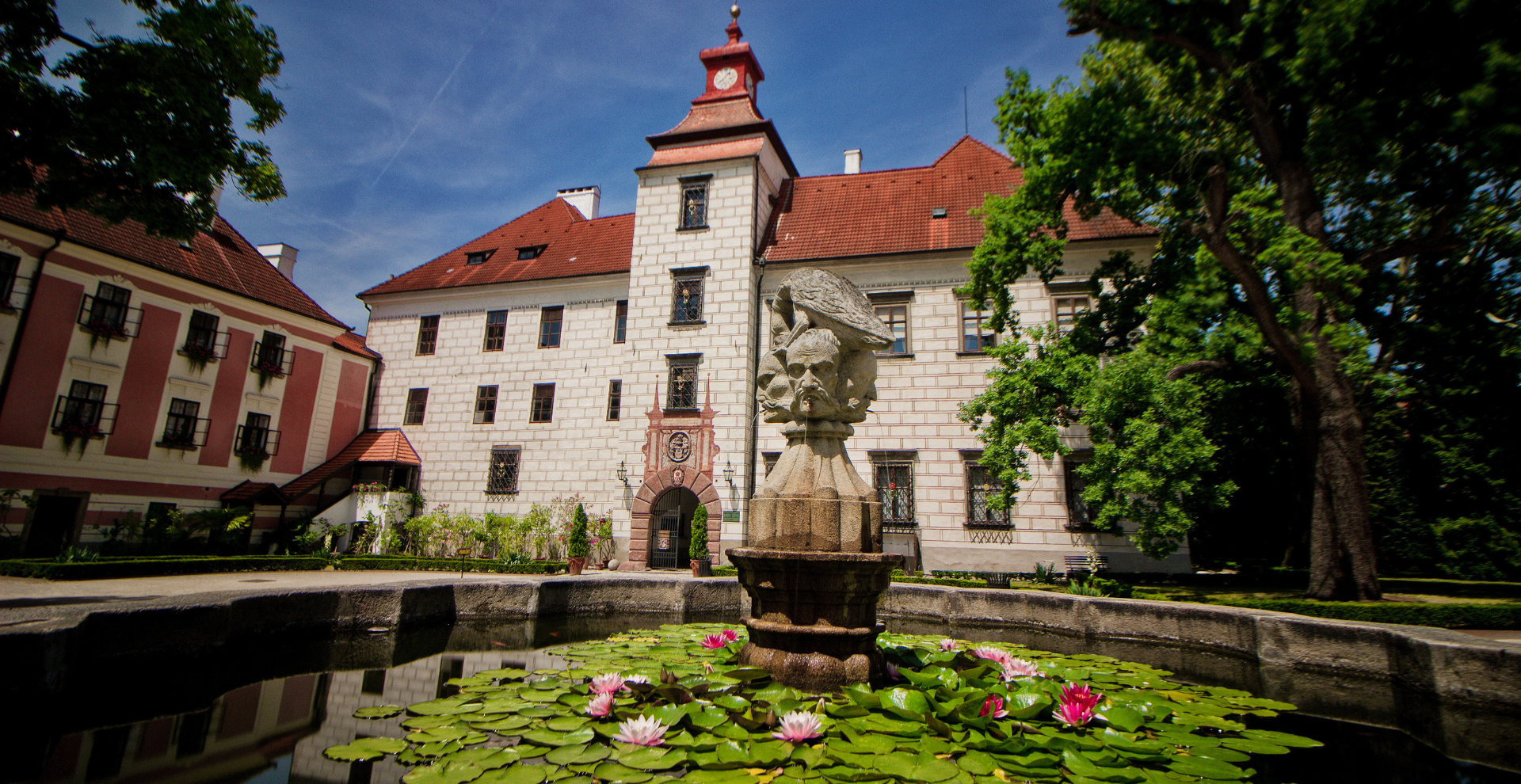 Historic courtyard with a red-tiled roof building, central clock tower, and a stone fountain amid water lilies.