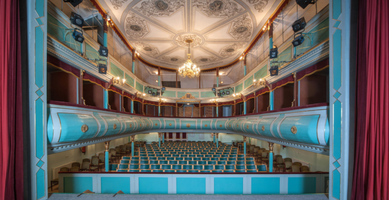 Interior of a historic theater with turquoise balconies, red velvet curtains, and a crystal chandelier.