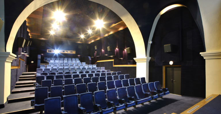 Empty theater auditorium with blue upholstered seats and cream arches, bright stage lights overhead.
