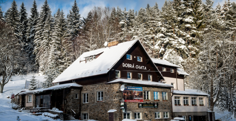 Snowy wooden lodge named 'Dobrá Chata' surrounded by a snow-covered evergreen forest under a blue sky in winter.