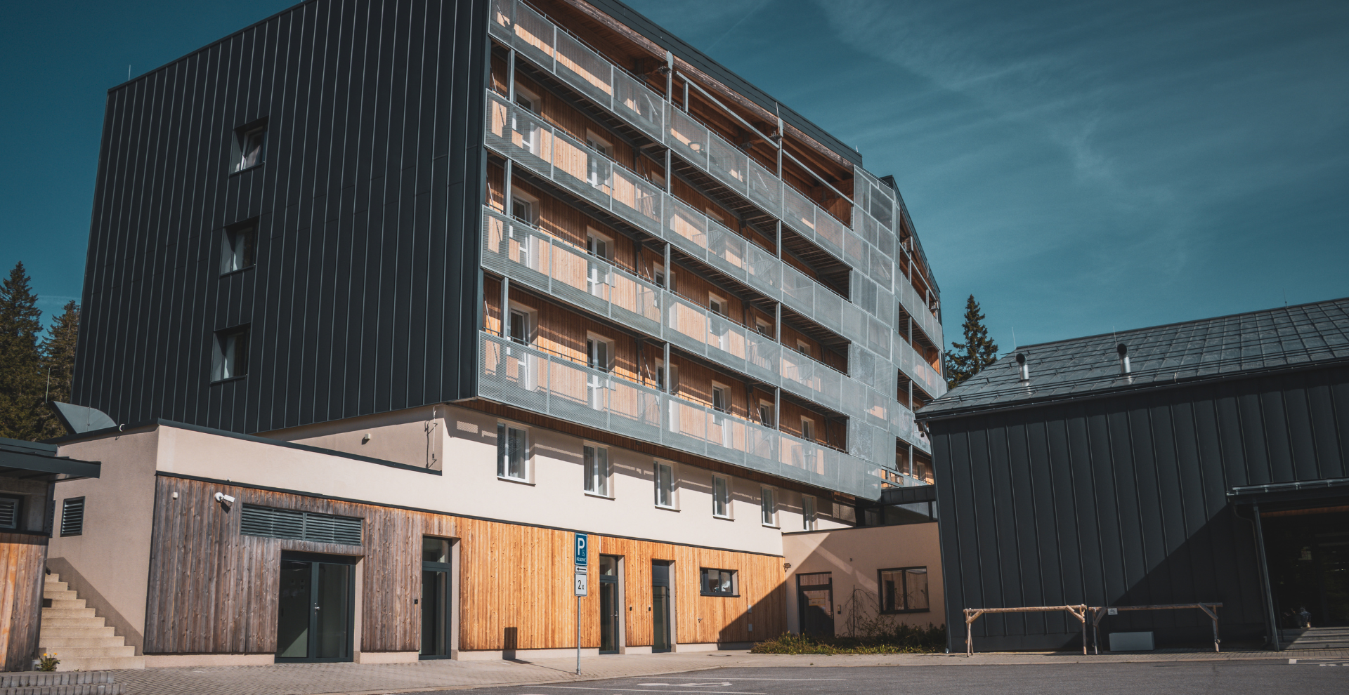 Modern multi-story residential building with wood and metal façade, long balconies with mesh railings, set against a blue sky.