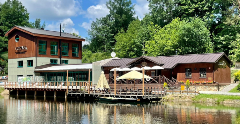 Lakeside restaurant with a wooden building and a docked deck over calm water, white umbrellas, and surrounding trees.