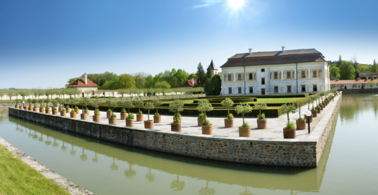 Sunlit historic manor with a symmetrical white facade, red roof, and formal terraced gardens edged by a moat.