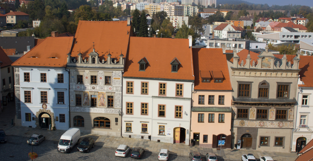 A row of historic buildings with orange tiled roofs along a cobblestone square; parked cars line the street and a museum is visible on the right.