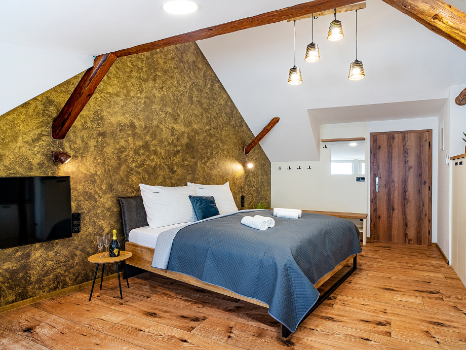 Hotel-style bedroom with a wooden beam ceiling, king bed, blue-gray quilt, white pillows, and a wall-mounted TV on the textured wall.