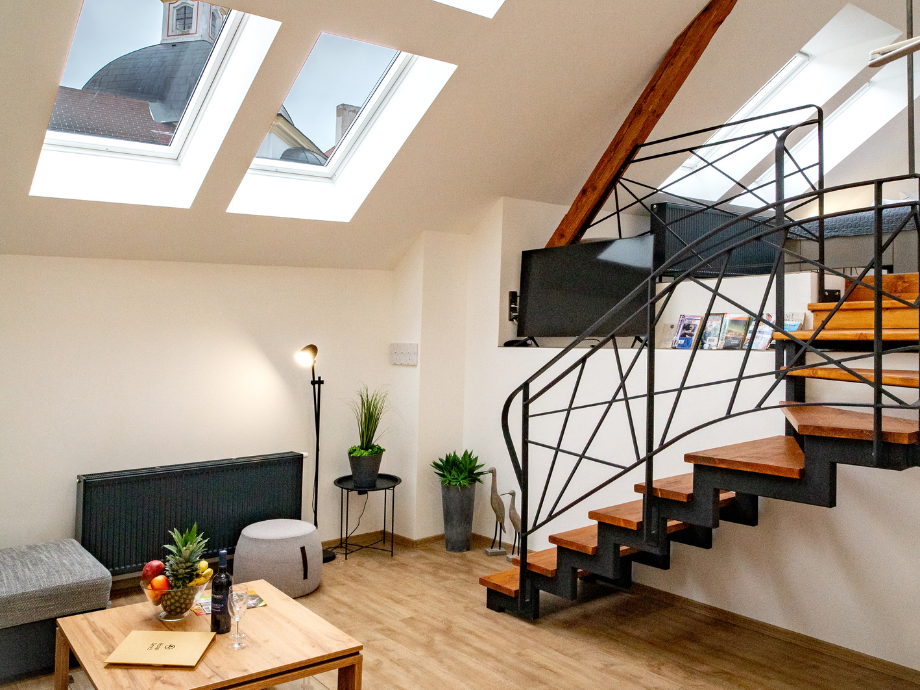 Loft interior with large skylights, a wooden staircase and black metal railing, plus a cozy seating area with plants and a coffee table.