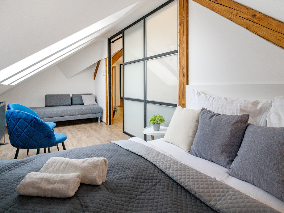 Attic bedroom with a gray quilted bed, blue chairs, a gray sofa, skylights, and a frosted glass divider framed by wood beams.