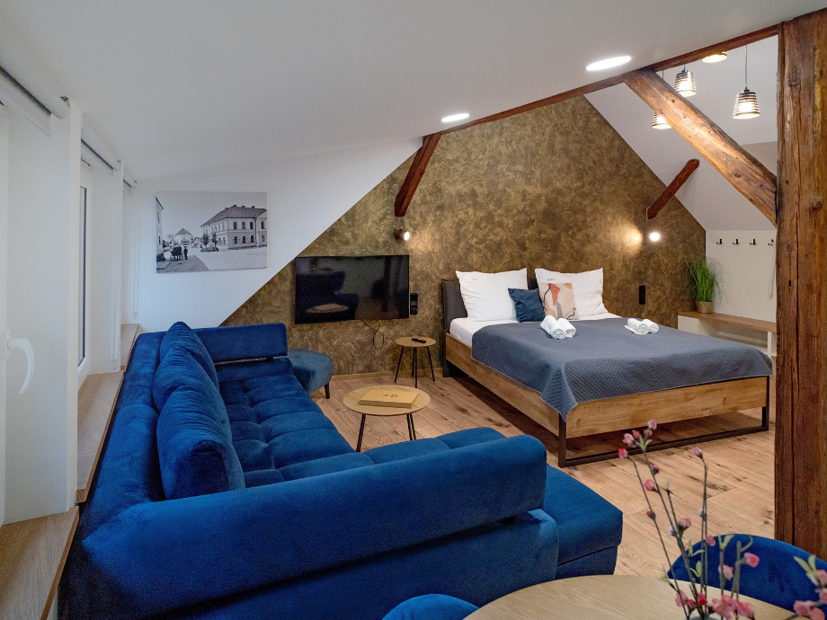 Loft-style hotel suite with a blue sectional sofa, round coffee tables, and a wooden bed under exposed beams.