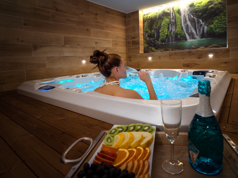 Woman relaxing in a white hot tub with blue-lit water, wooden spa walls, and a framed waterfall mural above. A tray of fruit, a champagne bottle, and a glass sit in the foreground.