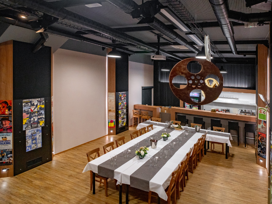 Long dining table set for a formal dinner in an event space, with flower centerpieces, wine glasses, and a hanging vintage film reel above; bar area in back.