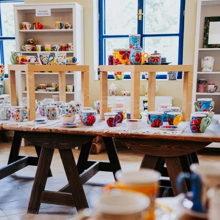Colorful handmade mugs and teacups arranged on a rustic wooden table in a bright pottery shop, shelves in the background.