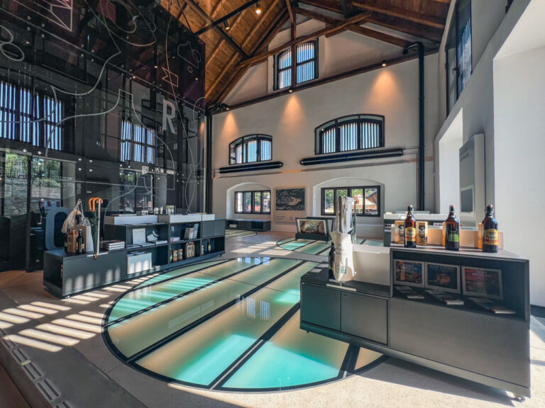 Industrial-style showroom with a curved glass-bottom bowling lane, dark shelving, and a bar displaying bottles.