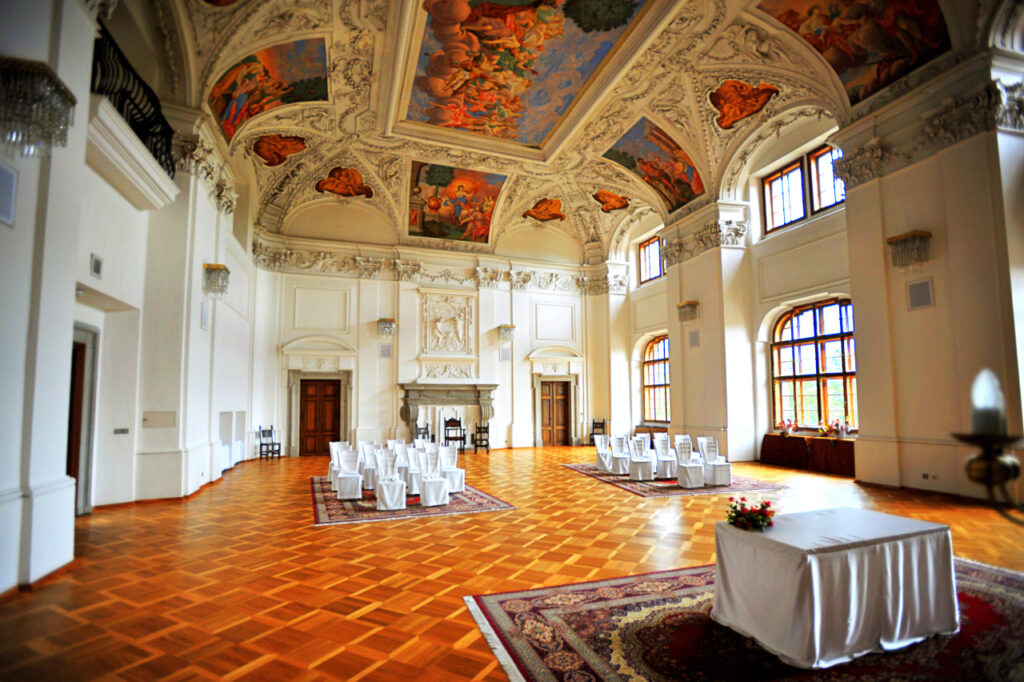Elegant ceremonial hall with a painted ceiling, tall arched windows, and rows of white-covered chairs facing a front dais with a flower centerpiece.